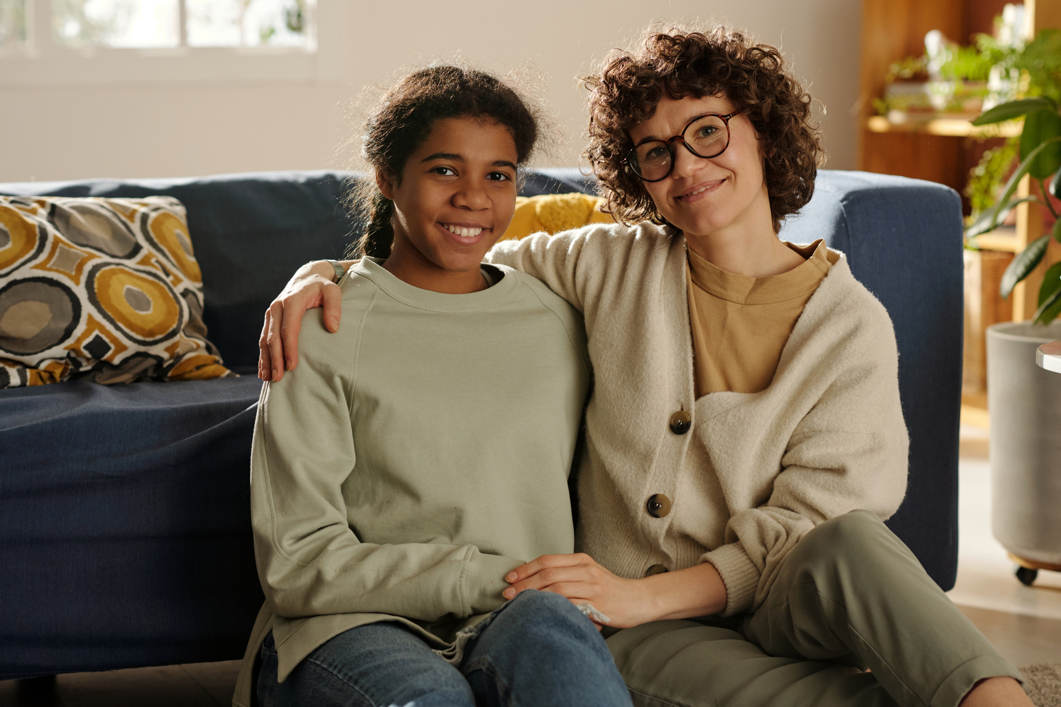 Portrait of young mom embracing her foster child while they are sitting on the floor and smiling at camera.